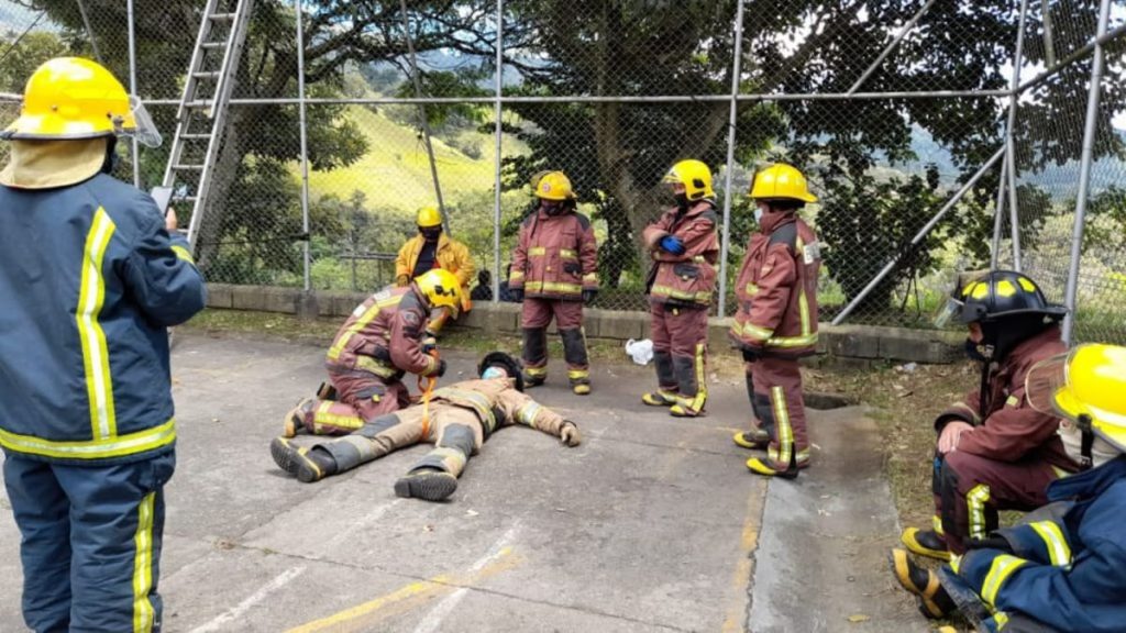 Bomberos con casco y traje de protección practican un rescate junto a un compañero tendido en el piso de una cancha