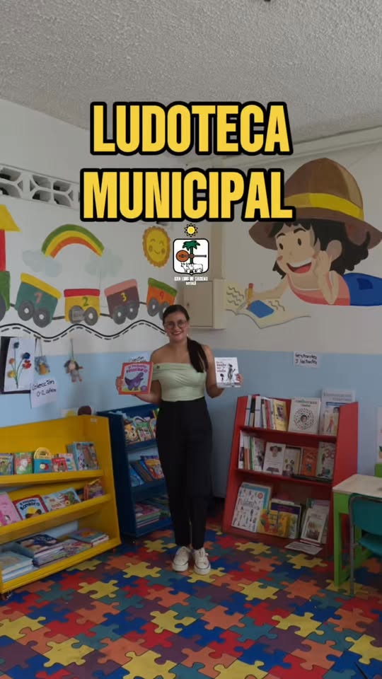 Una mujer muestra dos libros infantiles en la Ludoteca Municipal de San Luis de Gaceno, con murales, estantes de colores y piso de rompecabezas