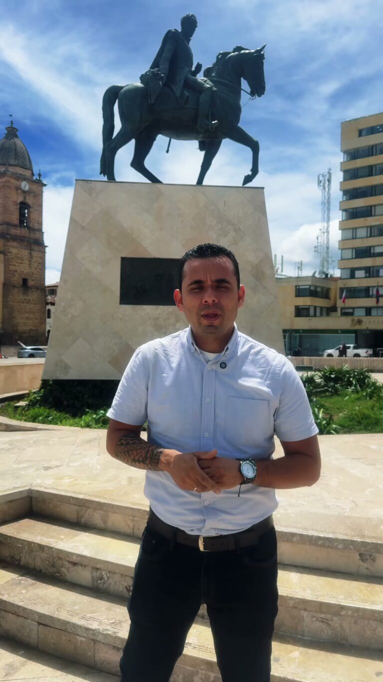 Un hombre de camisa clara invita a la elección de Asojuntas frente a una estatua ecuestre de Bolívar