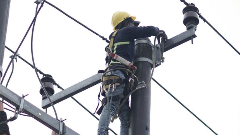 Un liniero con casco amarillo trabaja en lo alto de un poste de energía