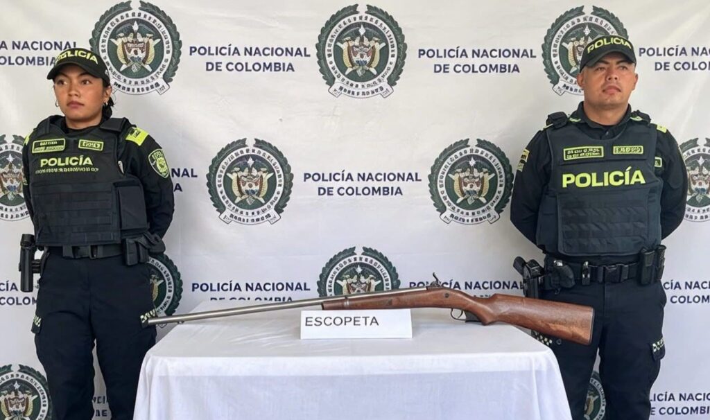 Dos policías de pie a lado y lado de una mesa con mantel blanco sobre la que está la escopeta hallada, con un letrero que dice «Escopeta»
