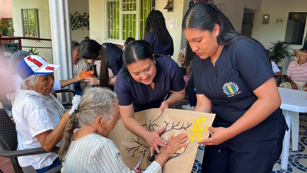 Una estudiante de Trabajo Social ayuda a una adulta mayor a poner su mano pintada en un árbol de papel