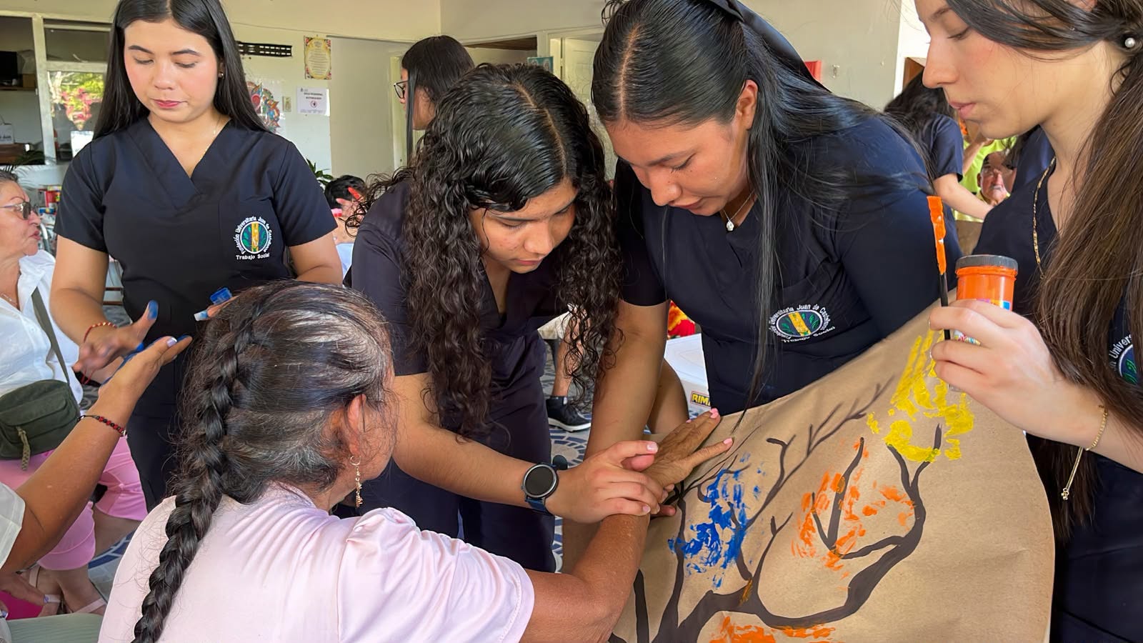 Estudiantes ayudan a una adulta mayor a pintar su mano en una cartelera