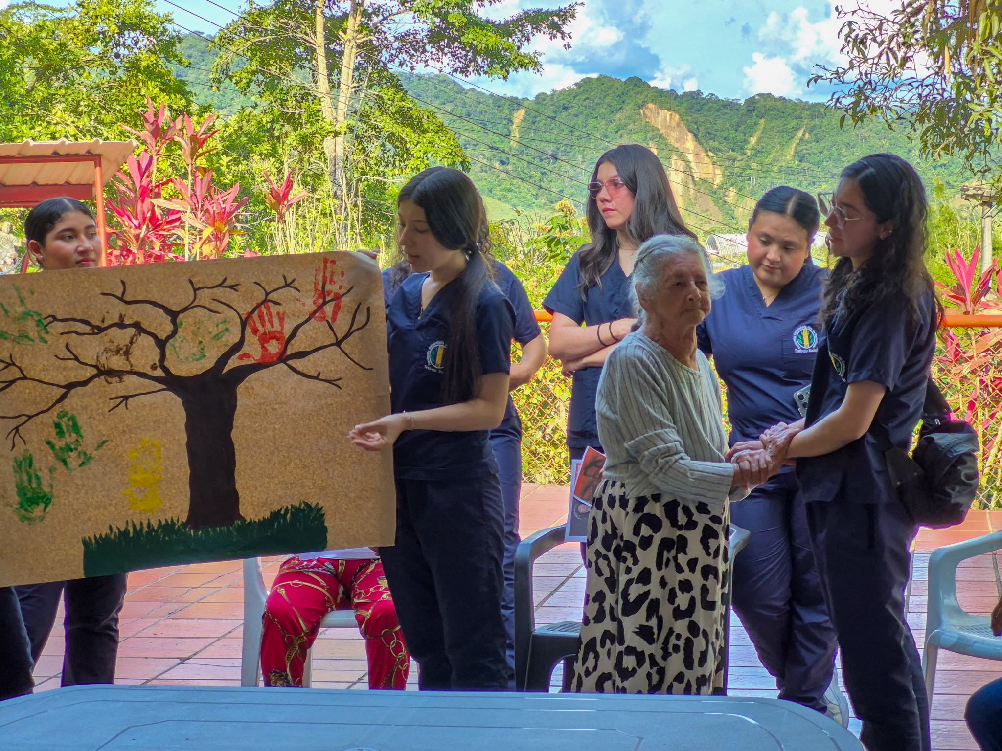Estudiantes muestran un árbol de papel con huellas de manos junto a una adulta mayor