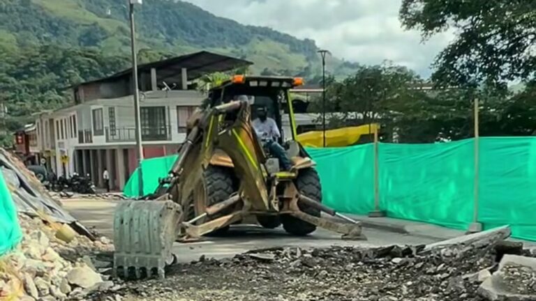 Retroexcavadora amarilla sobre una calle levantada de Santa María, entre polisombra verde, con casas y montañas al fondo